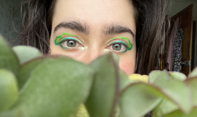 nera looking out from above a plant, they have green eyeliner